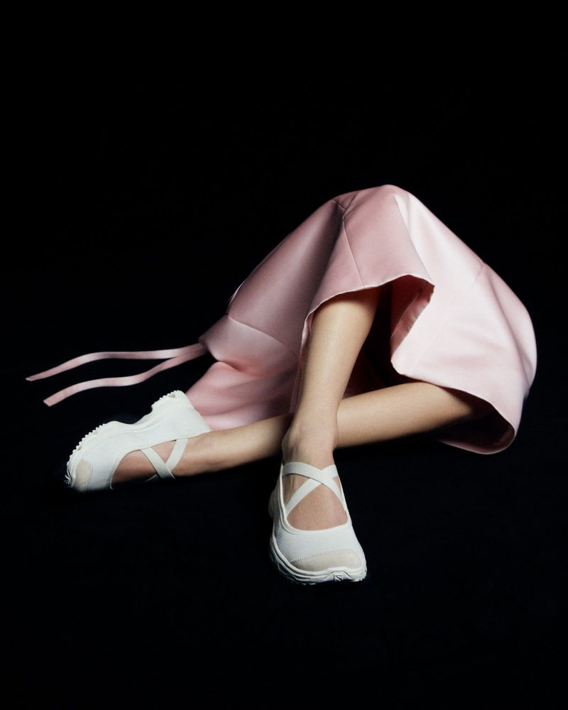 Abstract fashion shot of a pink dress with legs and white shoes extending from under it, against a stark black background.