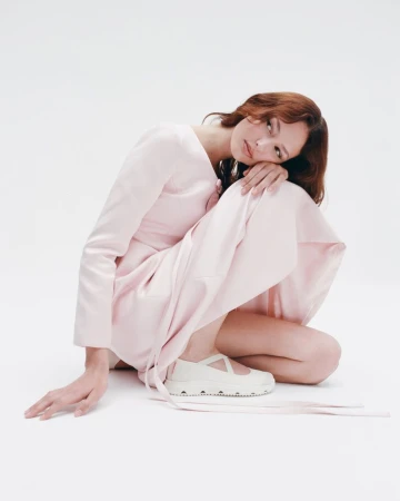 Fashion shoot: A woman in a light pink dress and cream Mary Jane shoes sits in a minimalist white studio, gazing softly at the camera.