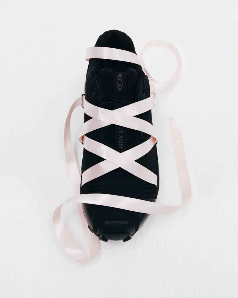Black shoe is wrapped with pale pink satin ribbons on a white surface, creating a modern and abstract still life.