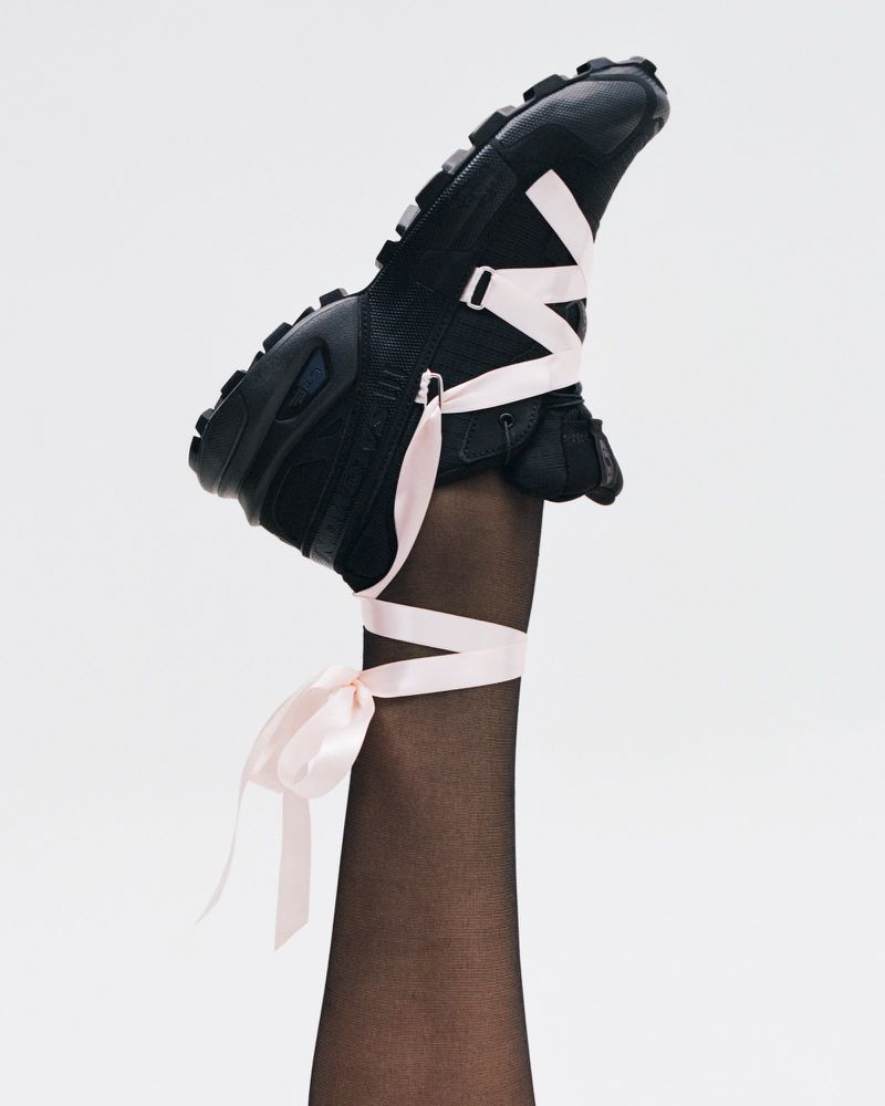 Leg wearing black hosiery and a black platform sneaker with pink ribbon laces pointing upward against a white backdrop in studio lighting.