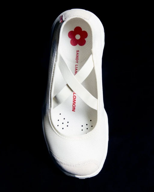Elevated view of a white Salomon x Sandy Liang sneaker with crossed straps on a black background, showcasing its ballet flat style.