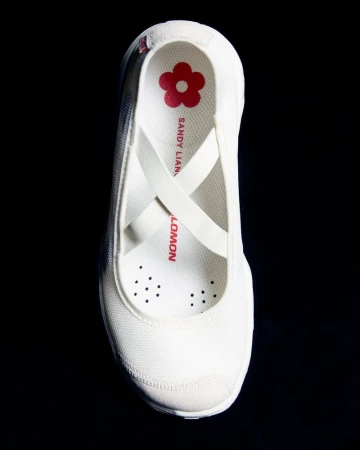 Elevated view of a white Salomon x Sandy Liang sneaker with crossed straps on a black background, showcasing its ballet flat style.
