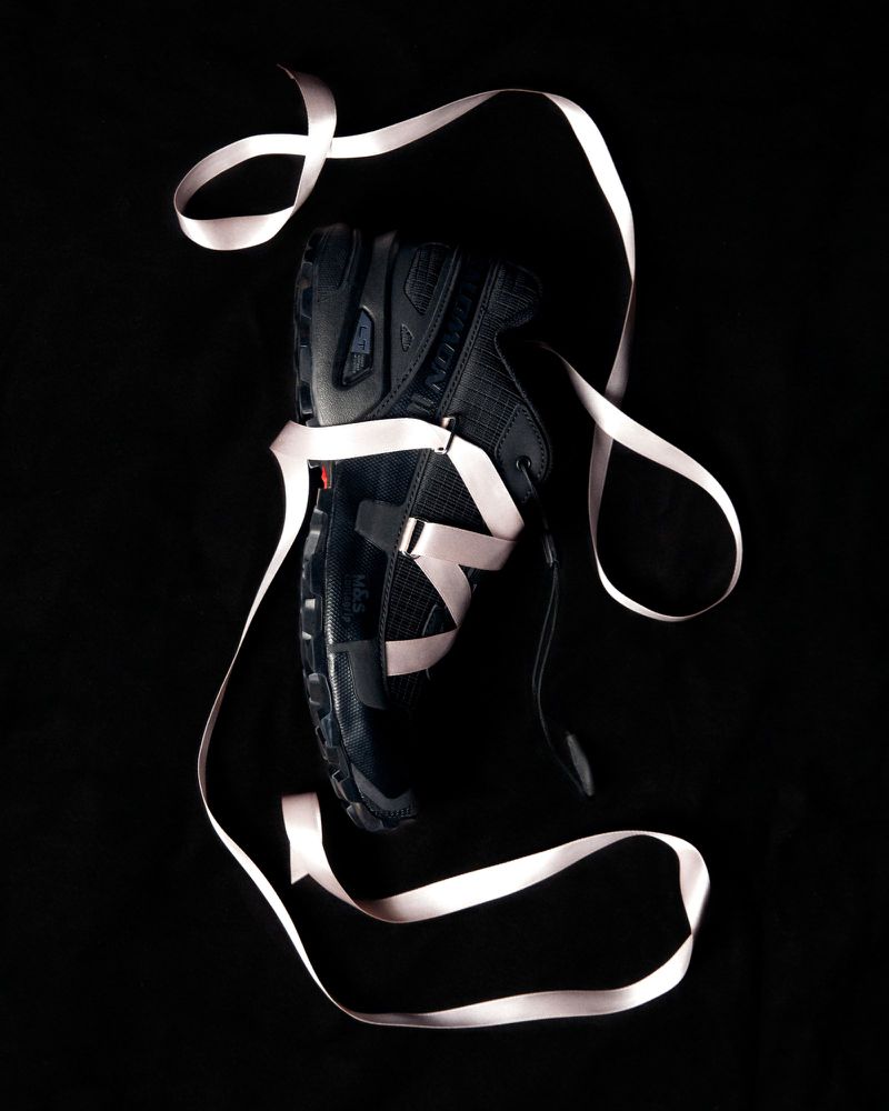 Black athletic shoe floats on a black background, wrapped with a curling pink ribbon. Dark, moody lighting and product shot.