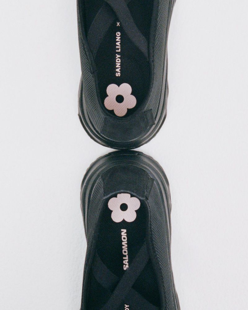 Two black Salomon x Sandy Liang Mary Jane shoes, shown close-up, reflecting off a glossy white background, featuring pink flower accents.