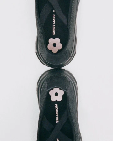 Two black Salomon x Sandy Liang Mary Jane shoes, shown close-up, reflecting off a glossy white background, featuring pink flower accents.