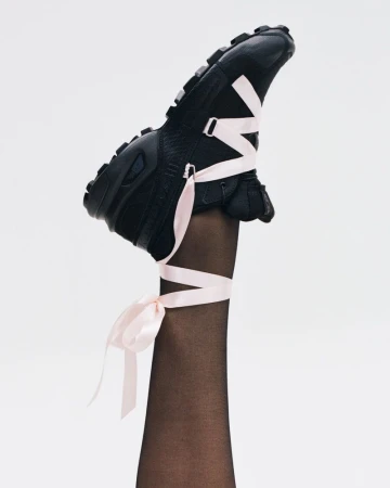 A raised leg in sheer black tights wearing a black trail sneaker laced with light pink satin ribbon tied in a bow at the ankle.
