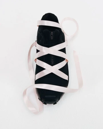 Top-down view of a black sneaker wrapped in crisscrossed pale pink satin ribbon against a clean white seamless studio backdrop.