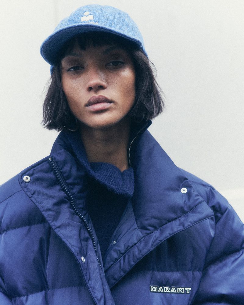 Portrait of a beautiful adult female model wearing a denim baseball cap and a navy blue puffer jacket with a neutral background.