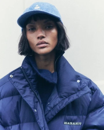 Portrait of a beautiful adult female model wearing a denim baseball cap and a navy blue puffer jacket with a neutral background.