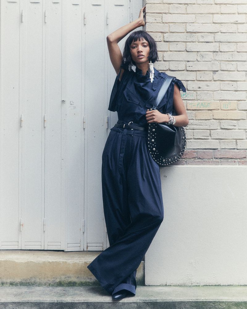 A stylish adult female poses in an outdoor setting, wearing wide-leg pants and a fashionable top, complemented by a studded shoulder bag.