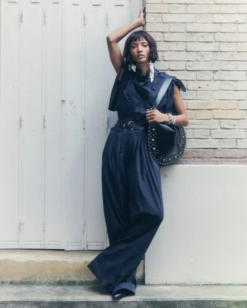 A stylish adult female poses in an outdoor setting, wearing wide-leg pants and a fashionable top, complemented by a studded shoulder bag.