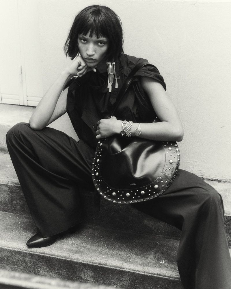 Woman in black outfit and shoulder bag with metal studs posing on stairs, black and white fashion shot, accessories on wrist, indoors.