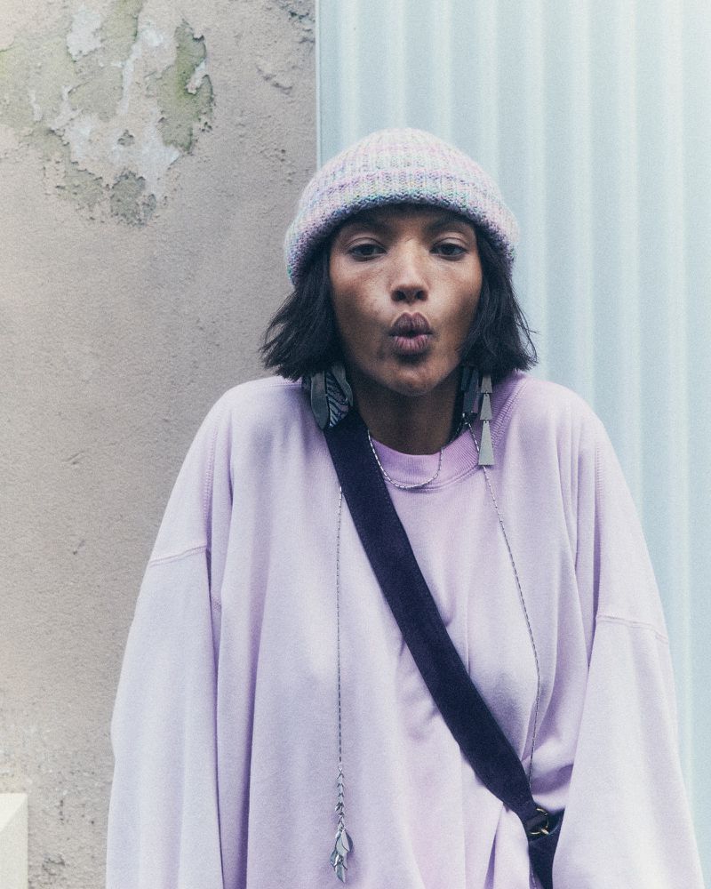 A person with dark hair wearing a knit hat and a lavender sweatshirt puckers their lips while standing in front of a textured wall.