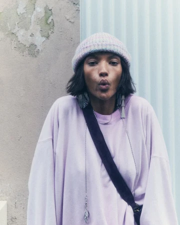 A person with dark hair wearing a knit hat and a lavender sweatshirt puckers their lips while standing in front of a textured wall.