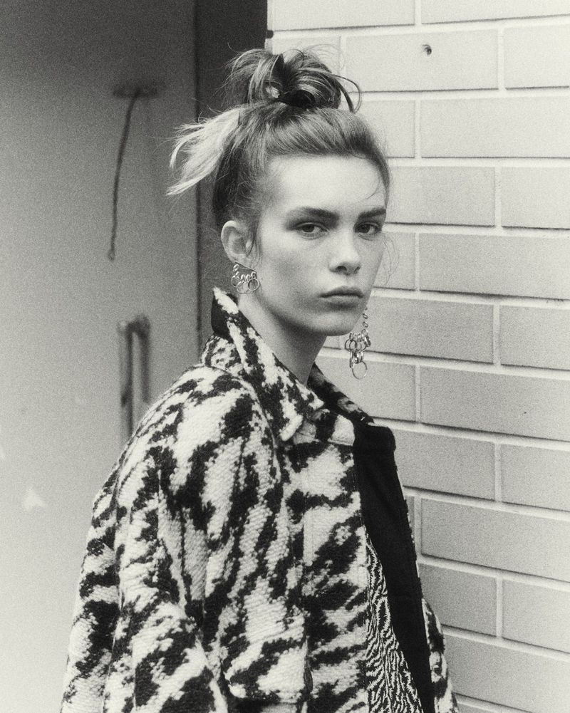 Black and white portrait of a young adult female model wearing earrings and a patterned jacket, standing against a brick wall.