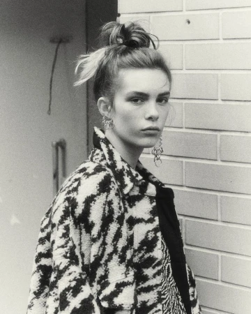 Black and white portrait of a young adult female model wearing earrings and a patterned jacket, standing against a brick wall.