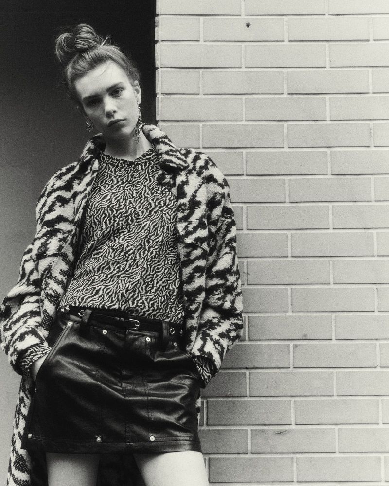 Young adult female poses in leather skirt and printed top leaning against a brick wall, wearing a patterned jacket in studio lighting.