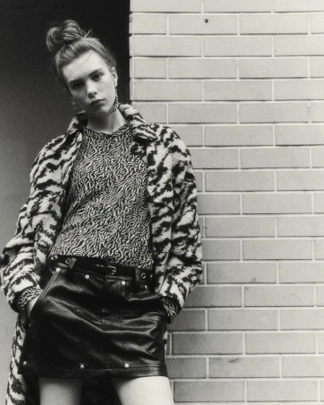 Young adult female poses in leather skirt and printed top leaning against a brick wall, wearing a patterned jacket in studio lighting.