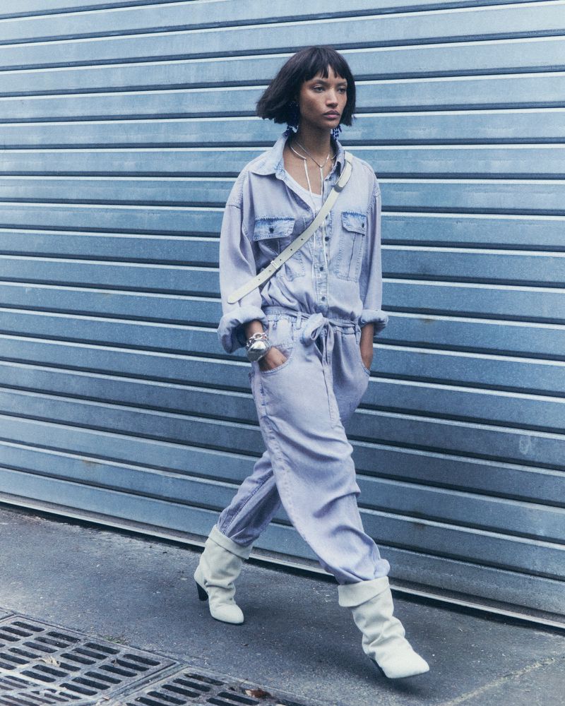 A stylish adult female strides confidently in a denim jumpsuit and white boots against a blue corrugated metal wall backdrop outdoors.