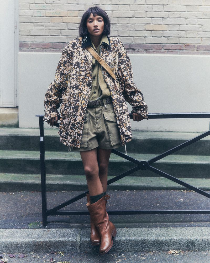 Adult female fashion model wears a patterned jacket, shirt, shorts, and boots outdoors against a brick wall and metal railing.