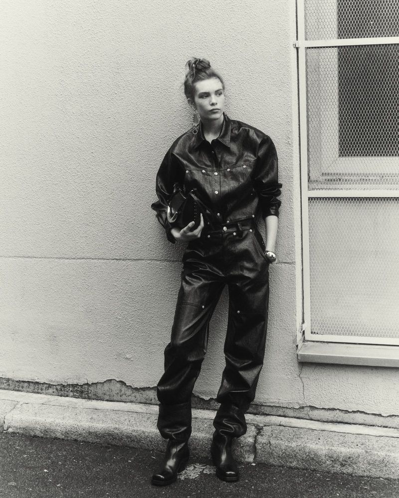 Model Freya Nutter wearing a black leather jumpsuit and boots, leaning against an exterior wall for Isabel Marant.