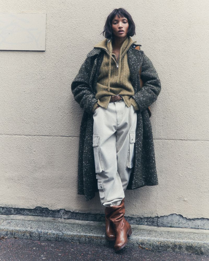 A woman with short brown hair in a brown and grey coat, yellow sweater, and white cargo pants leans against a wall outside.