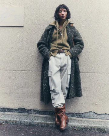 A woman with short brown hair in a brown and grey coat, yellow sweater, and white cargo pants leans against a wall outside.
