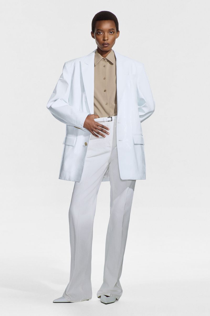A stylish, full-length shot features a Black female fashion model in an oversized white blazer, beige shirt, and white pants against a white backdrop.