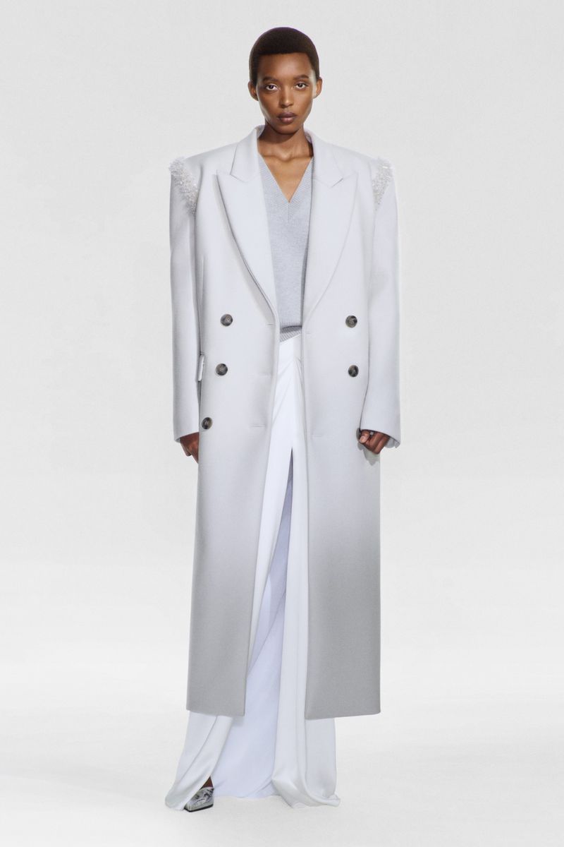 Elegant Black female fashion model in long, grey double-breasted coat and white skirt in a brightly lit photo studio setting.