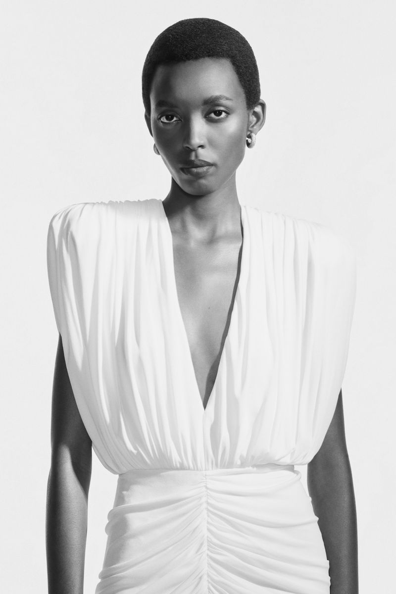 Black and white portrait of a Black woman in a white dress with a plunging neckline and gathered fabric, set against a neutral backdrop.