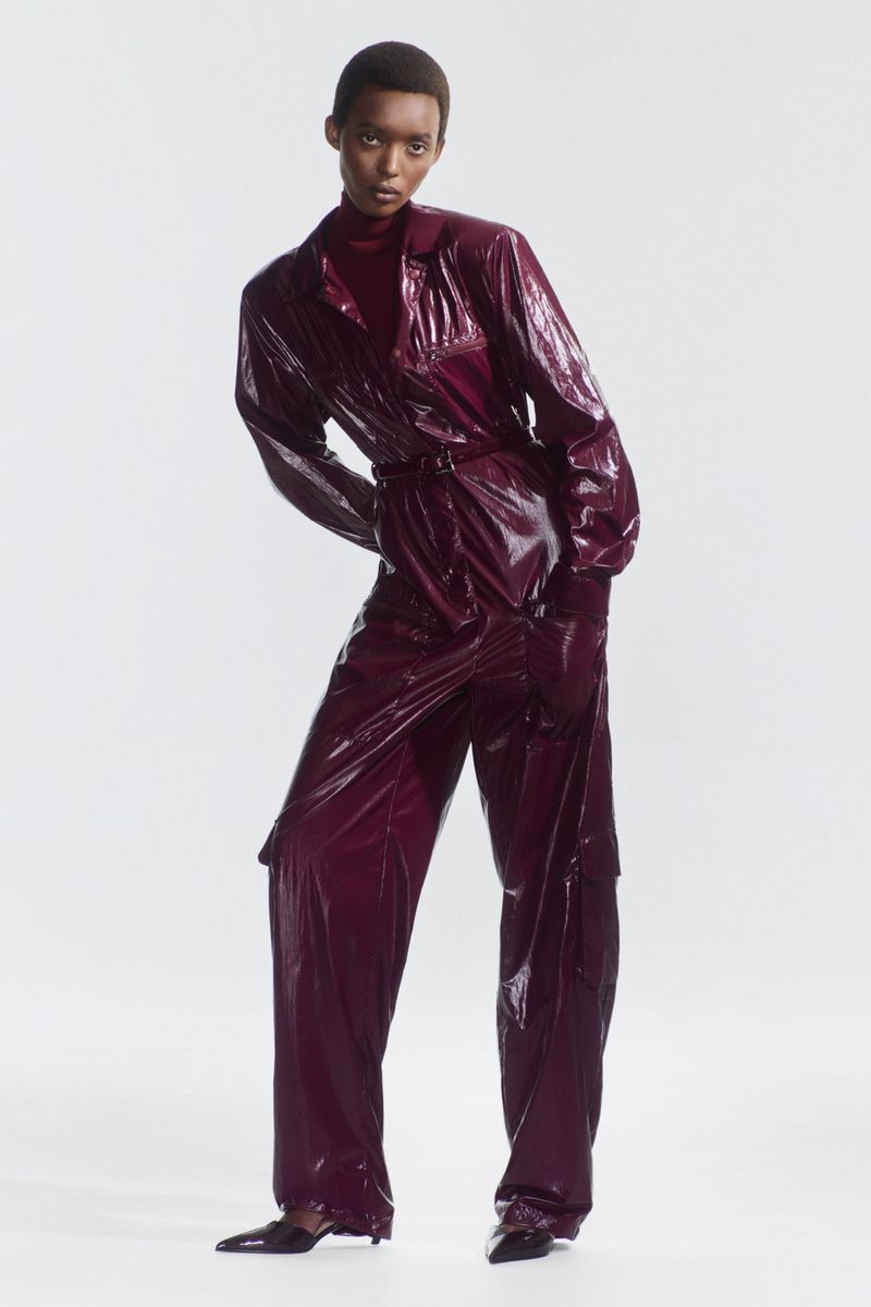An African American woman is posing in a burgundy, patent leather jumpsuit with black heels in a studio with a neutral background.