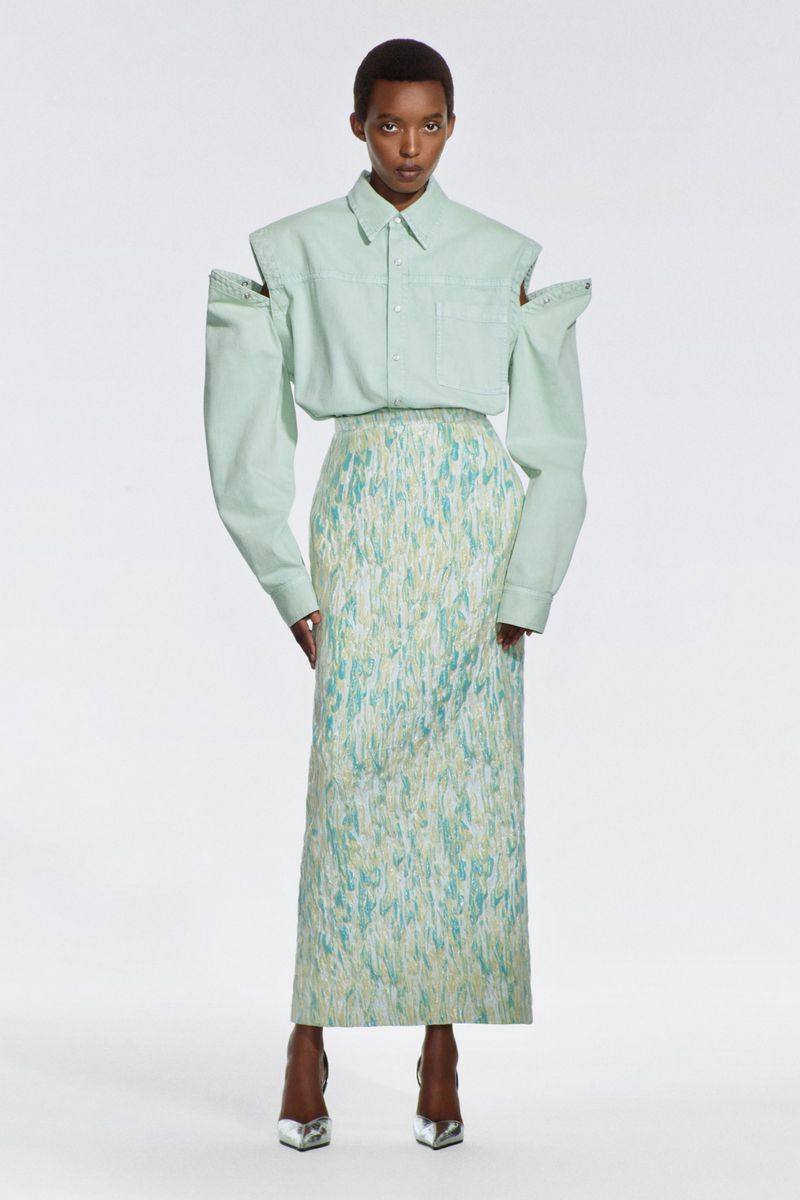 A single adult female fashion model poses in a light green shirt with shoulder cutouts and a long, patterned skirt in a white studio.