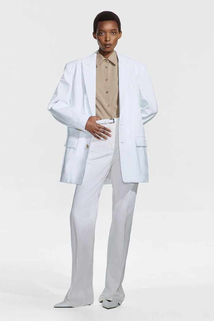 Model wearing an oversized white blazer, beige button-down shirt, and matching white trousers against a plain white studio backdrop.
