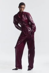 Model Claudia Ishimwe wearing a glossy burgundy belted jumpsuit and pointed shoes against a white background for Del Core Resort 2025.