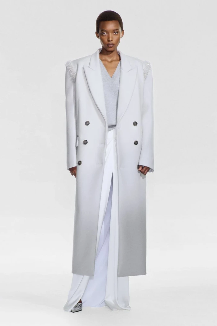 Model Claudia Ishimwe wearing a long double-breasted ombre coat and white wide-leg trousers against a white seamless background.