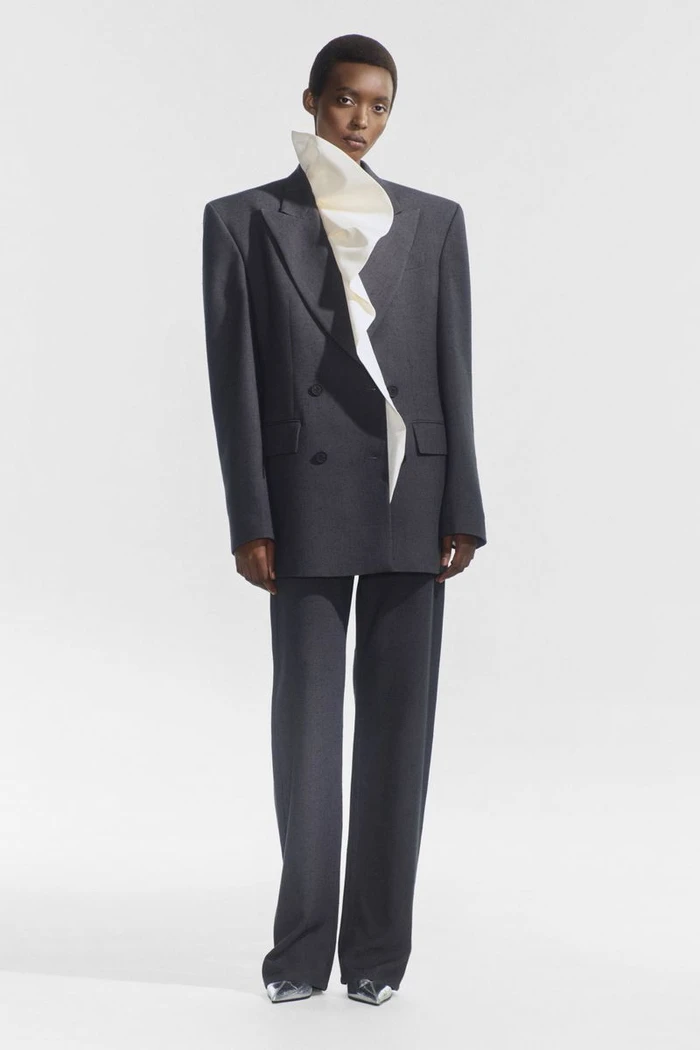 Model wearing an oversized charcoal grey tailored suit with a sculptural white ruffled collar against a bright white studio backdrop.