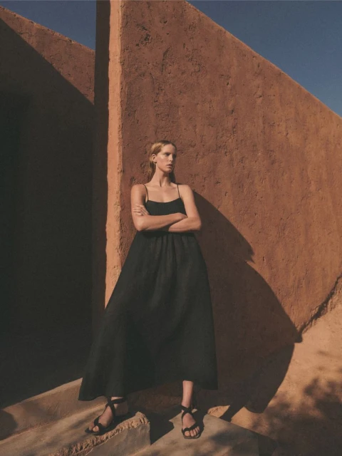 A slender woman in a black dress stands confidently in front of an earth-toned wall, bathed in warm light and casting soft shadows.