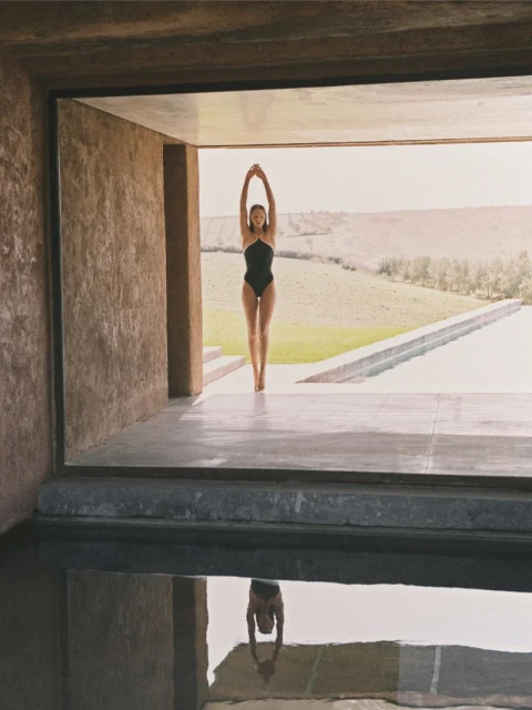 Young woman in a black swimsuit stands in a doorway of a modern building, reflected in the pool below with a green landscape backdrop.