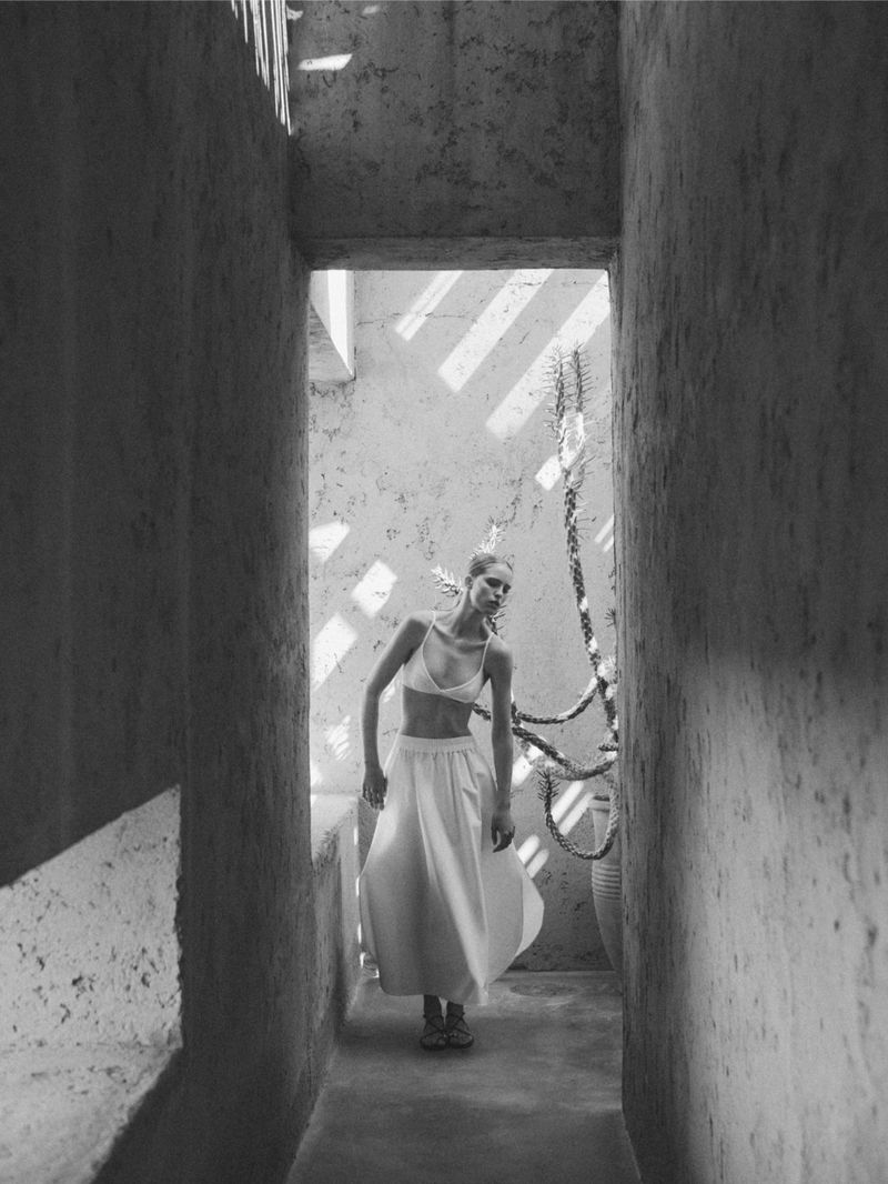 A woman in a white bra top and long skirt walks toward the camera through a passageway with light and shadow.