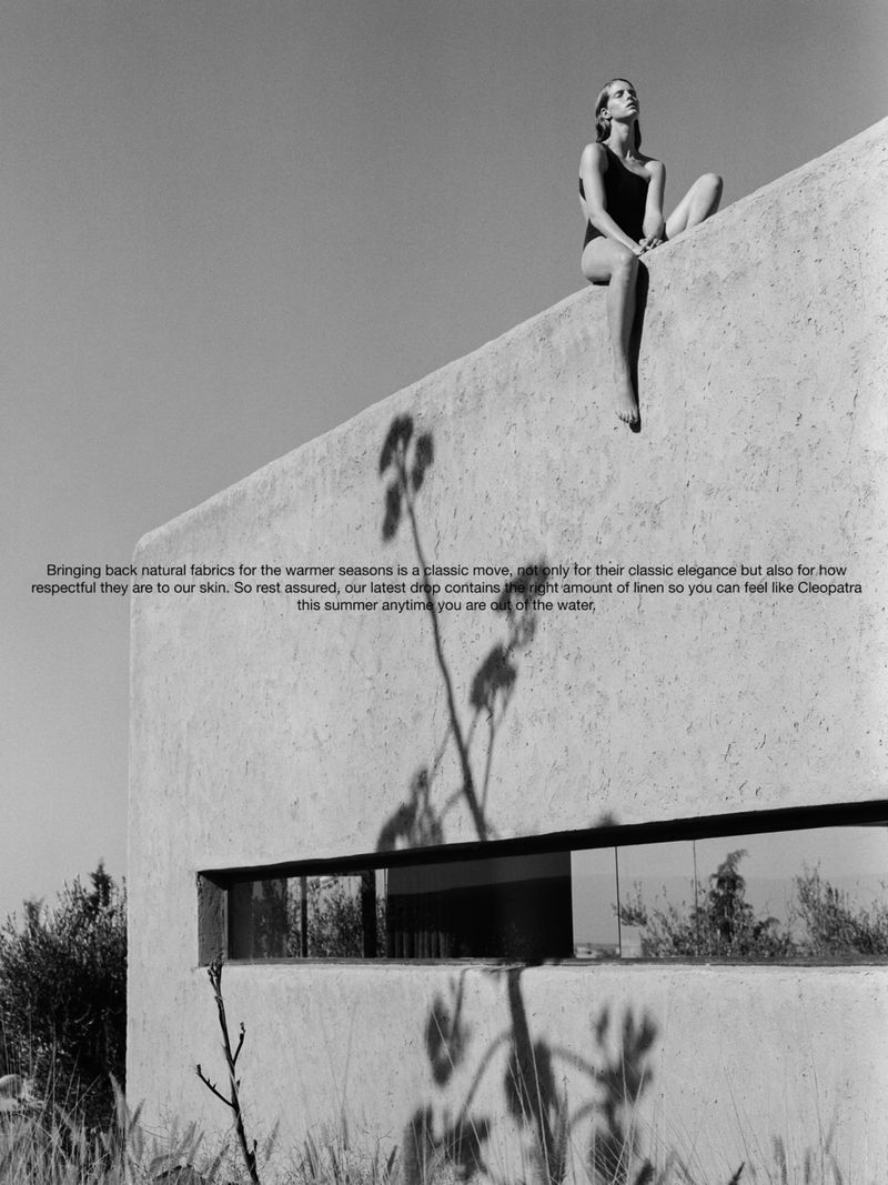 A woman in a swimsuit sits atop a concrete building against a sunny sky in a black and white fashion shot with architectural elements.