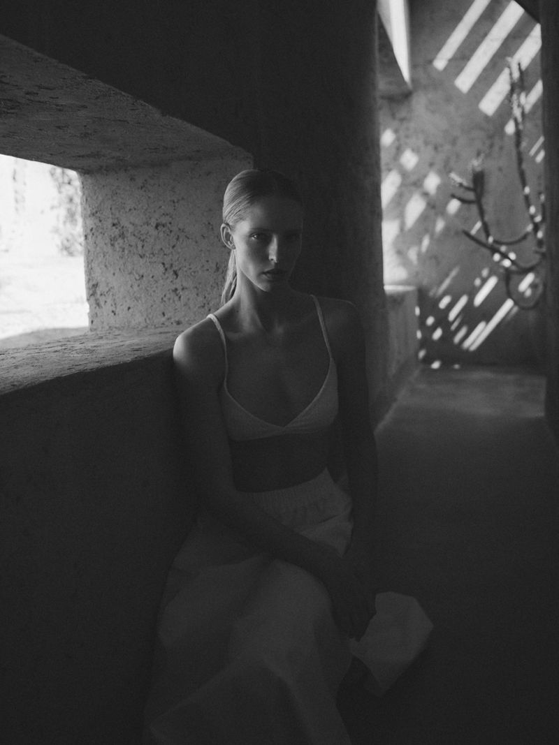 Black and white portrait of Abby Champion seated in swimwear in a shaded stone corridor with patterned sun shadows.