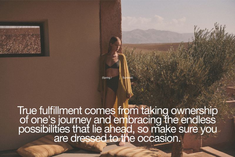 A woman stands outdoors in a yellow robe and swimsuit against a backdrop of an arid landscape with mountains under a bright sky.