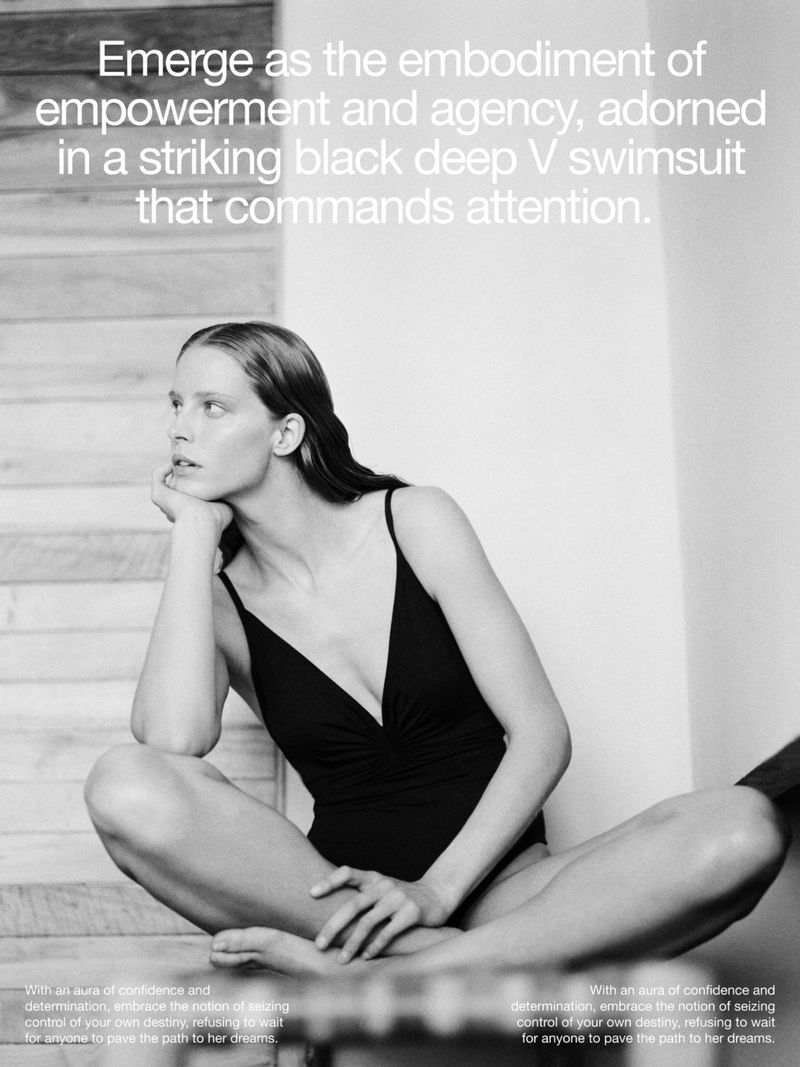 Black and white fashion shot of a slender, fair-skinned woman in a black v-neck swimsuit sitting cross-legged on a wooden staircase indoors.