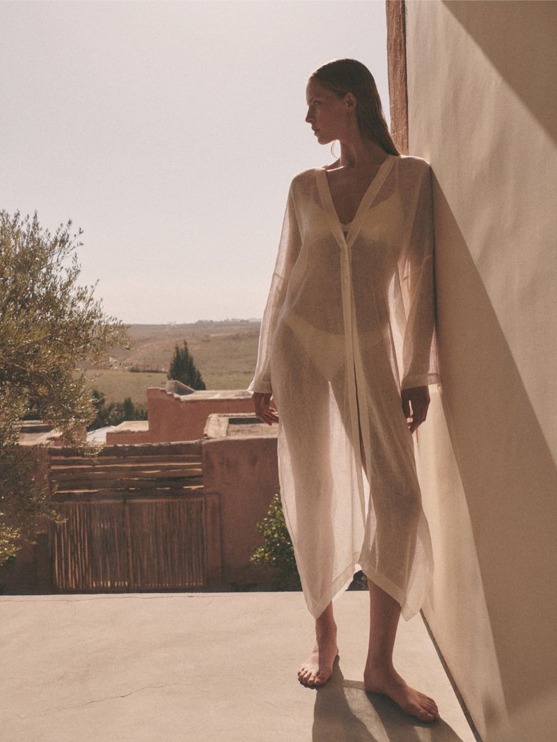 A woman in a sheer white robe stands on a balcony with a scenic landscape view, bathed in warm sunlight, creating a serene atmosphere.