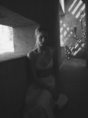 Black and white portrait of Abby Champion seated in swimwear in a shaded stone corridor with patterned sun shadows.