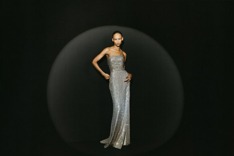 A woman in an elegant, floor-length silver gown poses against a dark background in studio lighting, creating a sophisticated fashion shot.