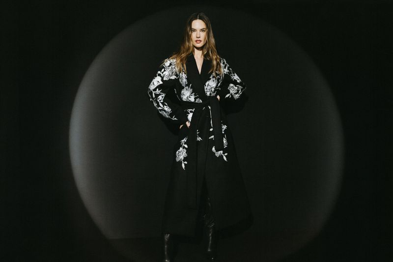 A full-length portrait of an adult woman with long hair posing in a black floral robe against a black backdrop with soft studio lighting.
