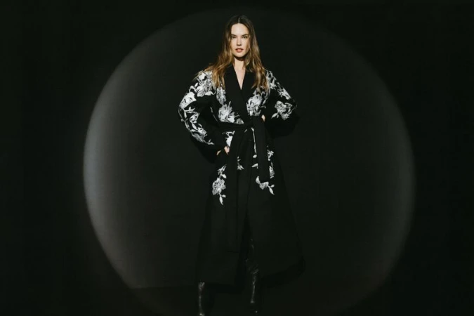 A full-length portrait of an adult woman with long hair posing in a black floral robe against a black backdrop with soft studio lighting.