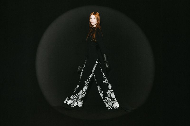 Fashionable woman poses in black outfit with floral patterned pants under a spotlight against a dark background in a studio setting.