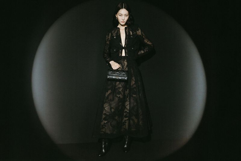 Elegant woman in black lace jacket and skirt holding a purse, posing in a dark photo studio with spotlight. Fashion photography.